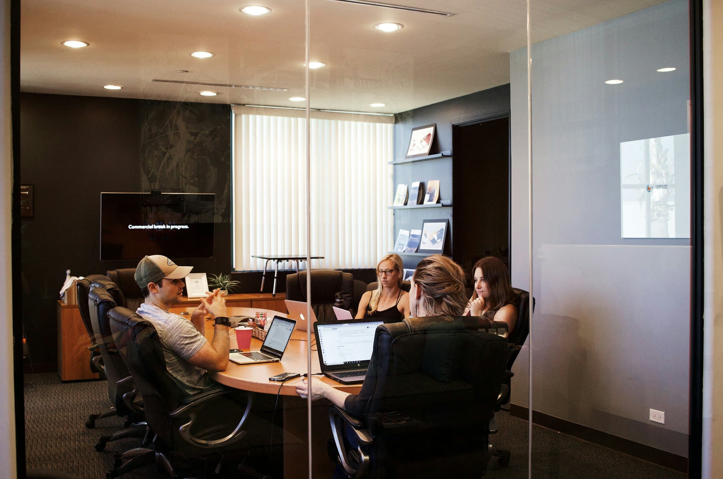 A small team working together with laptops around a table in a glass-walled meeting room